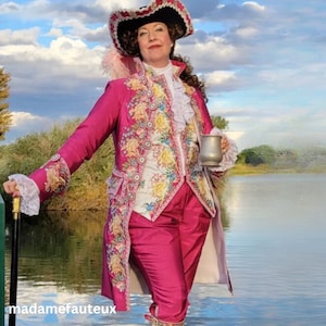 May include: A vibrant fuchsia coat with intricate floral embroidery and gold trim, worn over matching fuchsia trousers. The ensemble includes a white ruffled jabot, a tricorn hat with pink feathers, and a black cane with a gold handle. A metal tankard is held in one hand.