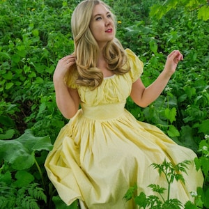 May include: A woman in a yellow dress with a ruffled neckline stands in a field of green plants. She is looking up and smiling.