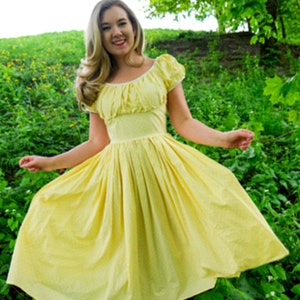 May include: A woman wearing a yellow dress with white polka dots and a white trim around the neckline. She is standing in a field of green grass and yellow flowers.
