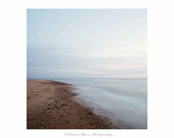 May include: A serene beach scene with a sandy shore and calm ocean under a pastel sky. The long exposure creates a blurred effect on the water, and the sand is dotted with small pebbles. The photograph is framed with a white border.