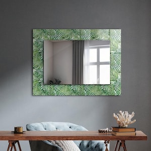 Puede incluir: Un escritorio de madera con un espejo con estampado de hojas verdes colgado encima. El escritorio está sobre una base de caballete y tiene un sillón azul delante. El escritorio está decorado con libros, una taza y una planta.