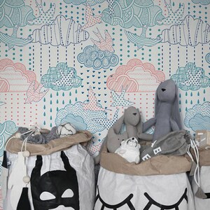 Puede incluir: Tres grandes bolsas de almacenamiento de lona blanca con dise&ntilde;os negros. La bolsa de la izquierda tiene un dise&ntilde;o de m&aacute;scara de Batman negro. La bolsa del medio tiene un dise&ntilde;o de ojo negro. La bolsa de la derecha tiene la palabra "TOYS" en letras negras. Las bolsas est&aacute;n llenas de animales de peluche y juguetes. El fondo es un papel tapiz blanco con un patr&oacute;n de nubes azules y rosas.