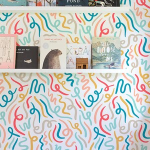 Pode incluir: Uma parede branca com um papel de parede de padrão abstrato colorido que apresenta um design repetitivo de linhas azul-esverdeadas, amarelas, rosadas e cinzas. Duas prateleiras brancas com livros estão montadas na parede. Os livros incluem títulos como "Maps", "Migrant", "Beyond the Pond", "Along a Long Road", "I Want My Hat Back", "Not My Hat", "This Moose Belongs to Me", e "Eiffel". Uma cesta de vime está no chão no canto inferior direito da imagem. O texto "Buy on kidsColoray Etsy Shop" está em um círculo preto na parte inferior da imagem.