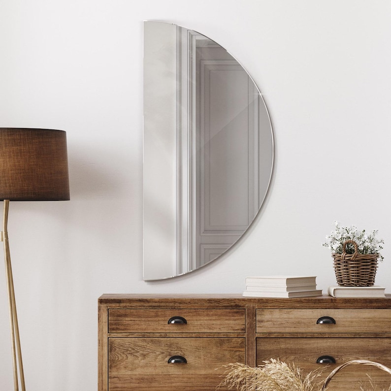 May include: A half-circle mirror with a silver frame hangs on a white wall above a wooden dresser with six drawers. A small potted plant sits on the dresser next to a stack of books.