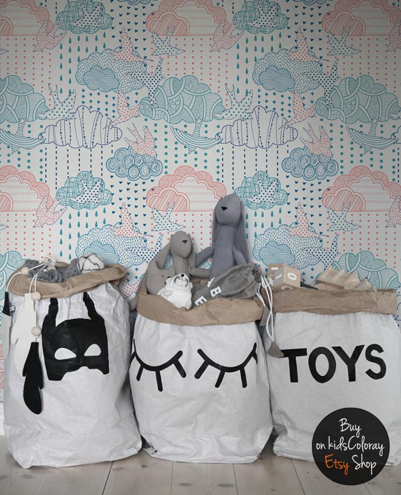 Puede incluir: Tres grandes bolsas de almacenamiento de lona blanca con dise&ntilde;os negros. La bolsa de la izquierda tiene un dise&ntilde;o de m&aacute;scara de Batman negro. La bolsa del medio tiene un dise&ntilde;o de ojo negro. La bolsa de la derecha tiene la palabra "TOYS" en letras negras. Las bolsas est&aacute;n llenas de animales de peluche y juguetes. El fondo es un papel tapiz blanco con un patr&oacute;n de nubes azules y rosas.