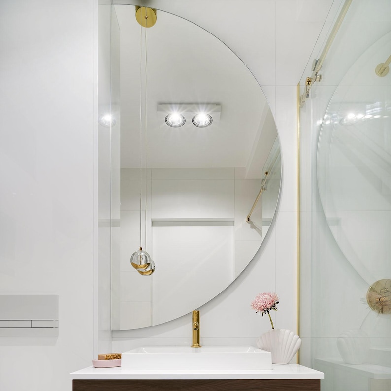 May include: A white bathroom with a half-round mirror, gold fixtures, and a white porcelain sink. A pink flower is in a white vase on the counter.