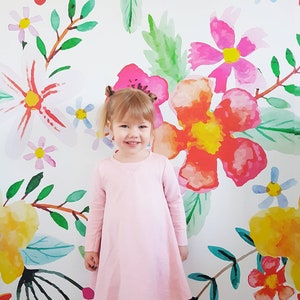 Puede incluir: Una niña pequeña con un vestido rosa se para frente a una pared blanca con un patrón floral. Las flores están pintadas en tonos de rosa, rojo, amarillo y verde.