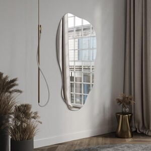 May include: A large, irregular-shaped mirror with a silver frame hangs on a white wall. The mirror reflects a window with a view of a city street.