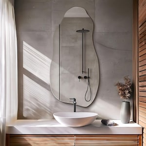May include: A modern bathroom with a white ceramic sink and a large, irregular-shaped mirror. The mirror reflects a shower with a black shower head and fixtures. The bathroom has a concrete wall and a wooden vanity.