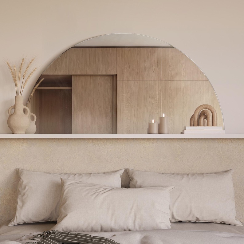 May include: A bedroom with a white headboard and a bed with white bedding. The headboard has a curved mirror above it, reflecting a wooden closet with sliding doors. There are two candles and a wooden sculpture on the shelf above the mirror.