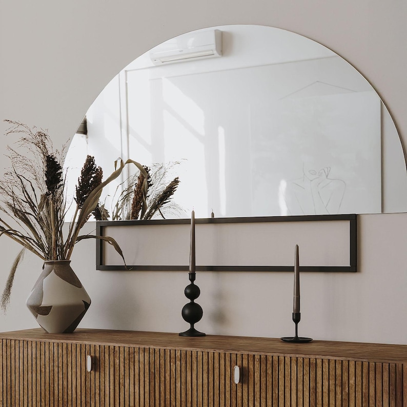 May include: A wooden cabinet with a black metal shelf above it. The cabinet has a wood grain pattern and two doors with oval handles. There are two black candle holders with black candles on the shelf. A vase with dried flowers is on the left side of the cabinet.