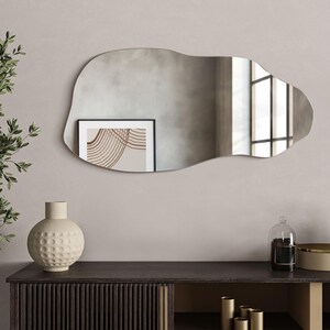 May include: A large, irregular-shaped mirror with a wavy, organic design. The mirror is hanging on a light gray wall above a dark wood dresser with a vase and other decorative items.