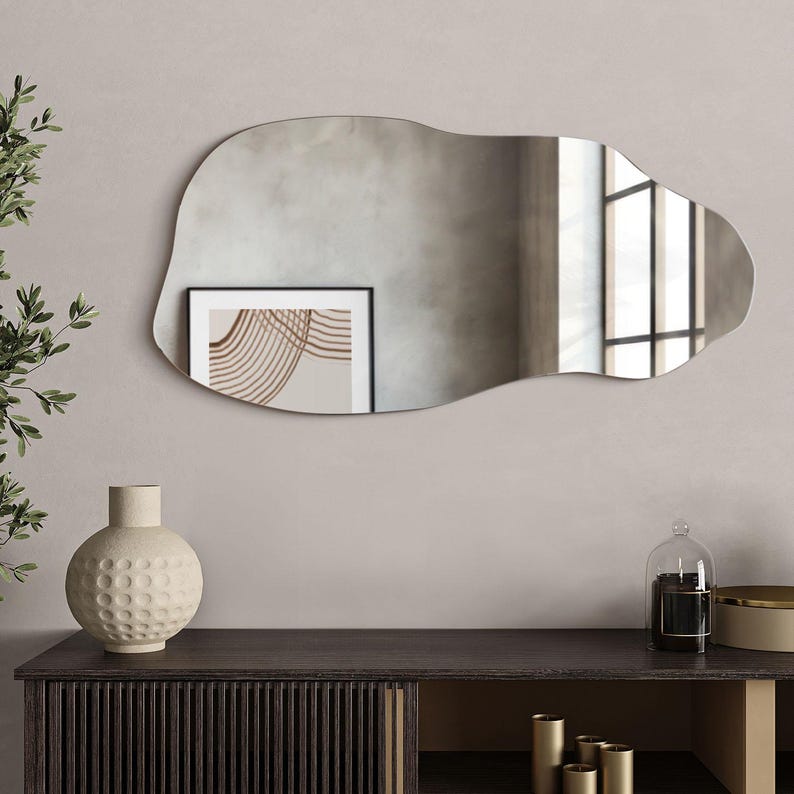 May include: A large, irregular-shaped mirror with a wavy, organic design. The mirror is hanging on a light gray wall above a dark wood dresser with a vase and other decorative items.