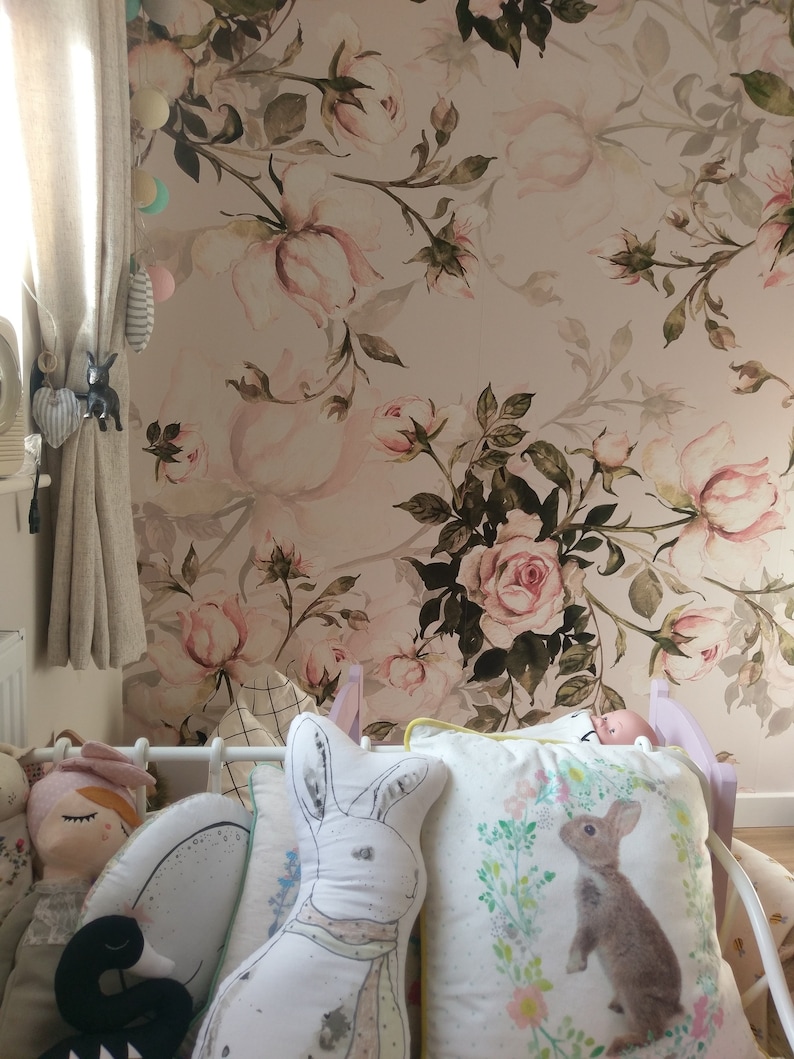 May include: A nursery room with a floral wallpaper featuring pink and green roses. The room has a crib with a white and gray bunny pillow, a white pillow with a brown bunny, and a doll with a pink dress.