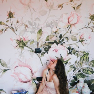 May include: A woman with long brown hair sits on a patterned rug in front of a wall with a floral mural. The mural features large pink roses with green leaves and stems. The woman is wearing a pink dress and is looking up at the mural.