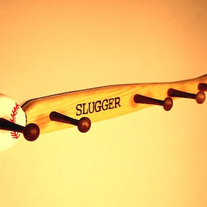 May include: Wooden baseball bat coat rack with five hooks. The bat is painted brown and has the word "SLUGGER" written on it. The end of the bat is shaped like a baseball with red stitching.