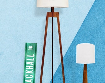 May include: A wooden tripod floor lamp with a white drum shade. The lamp is next to a smaller table lamp with a white shade. The background is a blue and light blue wall with a green sign that reads "BLACKHALL DR."