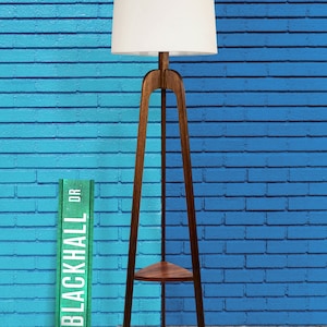 May include: A wooden floor lamp with a white lampshade and a shelf. The lamp is standing in front of a blue brick wall with a green sign that says "Blackhall Dr."