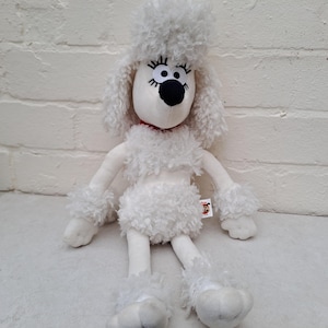 May include: A white plush poodle toy with a fluffy head, chest, and paws. The toy has a black nose, large eyes, and a red collar. The poodle is sitting on a white surface.