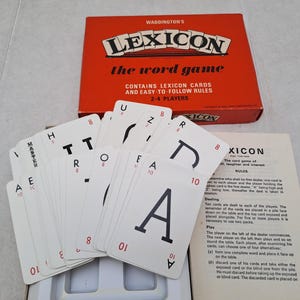 May include: A Waddington's Lexicon word game set featuring a red box with white text, a white instruction sheet, and a fan of white playing cards. Each card displays a large black letter, a small red letter in the corner, and a red number value.