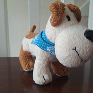 Può includere: Un giocattolo a forma di cane di peluche con corpo bianco, orecchie marroni e macchie marroni. Ha un naso nero, una sciarpa blu a pois e dettagli ricamati. Il giocattolo è progettato per assomigliare a un cucciolo amichevole.
