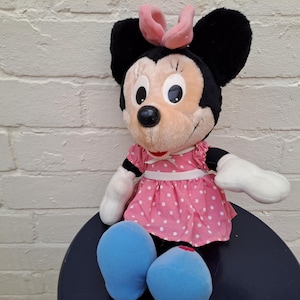 May include: A Minnie Mouse plush toy. The stuffed animal has black ears, a pink bow, and a pink dress with white polka dots. It has white gloves and blue shoes. The toy is sitting on a dark blue surface.