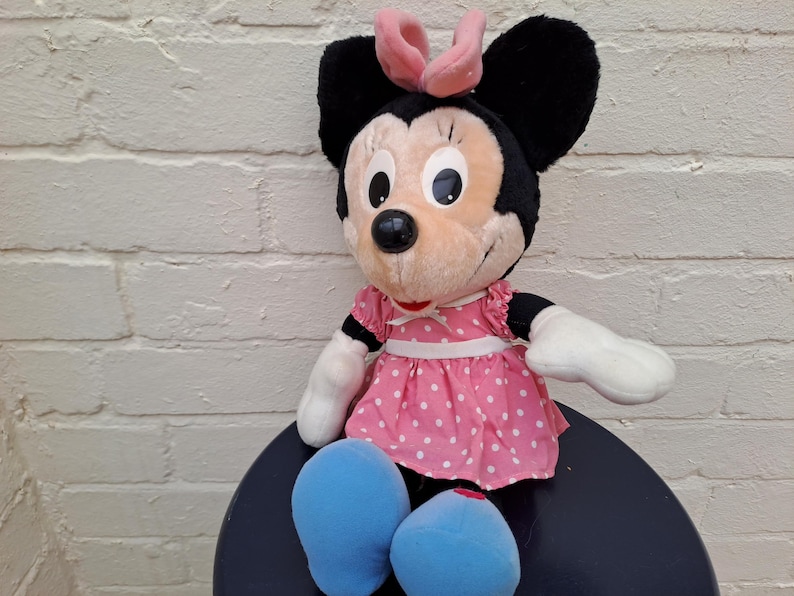 May include: A Minnie Mouse plush toy. The stuffed animal has black ears, a pink bow, and a pink dress with white polka dots. It has white gloves and blue shoes. The toy is sitting on a dark blue surface.