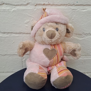May include: A plush teddy bear wearing a pink hat and outfit. The bear has a brown heart on its chest and striped accents. The bear's body is a mix of pink and tan colors, with brown paws.