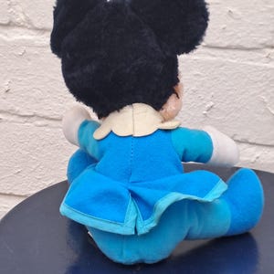 May include: A plush toy of a cartoon character, featuring a black head with large ears and a blue bow. The character wears a blue dress with a white collar and white gloves. The toy is sitting on a dark blue surface.