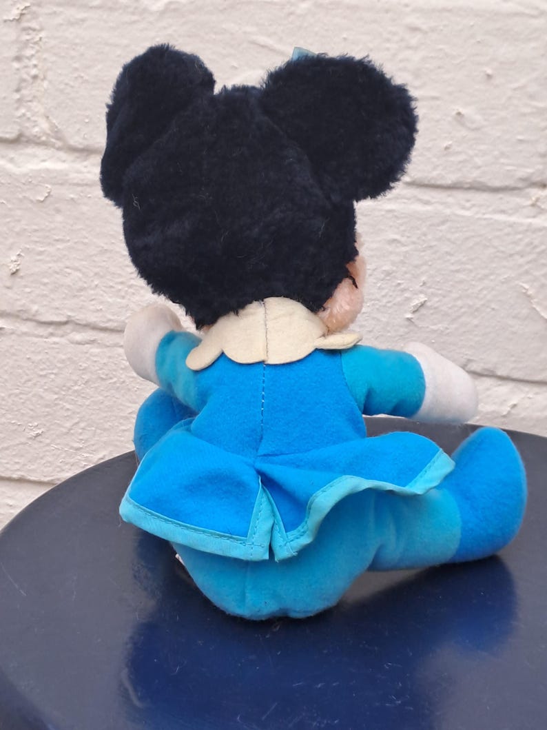 May include: A plush toy of a cartoon character, featuring a black head with large ears and a blue bow. The character wears a blue dress with a white collar and white gloves. The toy is sitting on a dark blue surface.
