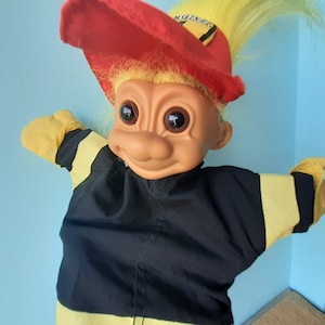 Puede incluir: Un muñeco troll de pelo amarillo que lleva un traje de bombero negro y amarillo con un sombrero rojo que dice "No Worries".