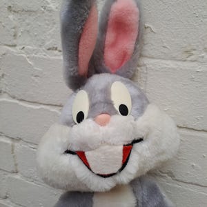 Può includere: Un peluche di Bugs Bunny. Il coniglio ha la pelliccia grigia con l'interno delle orecchie rosa e un muso bianco. Gli occhi sono neri e la bocca rossa. Il giocattolo è contro un muro di mattoni bianchi.