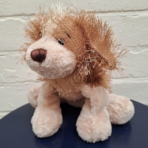 Puede incluir: Un juguete de peluche con forma de perro, con una combinación de colores crema y marrón claro. El perro tiene una cabeza y orejas esponjosas, una nariz marrón y patas suaves. El juguete está sobre una superficie azul oscuro.
