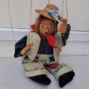 Könnte beinhalten: Eine Vintage-Marionettenpuppe, bekleidet mit einem karierten Hemd, einer beigen Weste und einer Hose aus Jute. Die Puppe hat einen Strohhut mit blauem Band, einen roten Schal und Holzzubehör. Die Puppe ist an Schnüren aufgehängt.