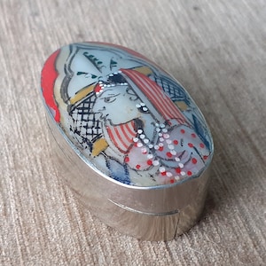 Pode incluir: Uma caixa de joias oval de prata com um desenho pintado de uma pessoa vestindo uma camisa listrada vermelha e branca e um chapéu branco com um padrão preto e branco. A pessoa está segurando um fio de contas brancas com pontos brancos.