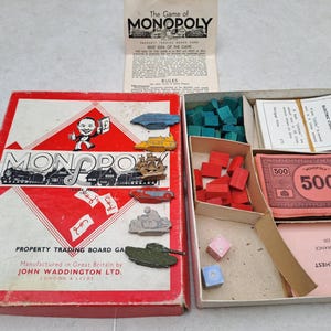 Könnte beinhalten: Ein klassisches Monopoly-Brettspielset mit einer rot-weißen Schachtel, dem ikonischen Charakter, Metallspielfiguren wie einem Schiff und einem Panzer, roten und grünen Holzhäusern, Spielgeld, Gemeinschaftskarten und einem Regelheft.