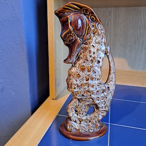 Könnte beinhalten: Eine braune und weiße Keramik-Seepferdchen-Figur mit strukturierter Oberfläche. Die Figur hat ein Loch an der Seite, was darauf hindeutet, dass es sich um eine Vase oder einen Krug handeln könnte.