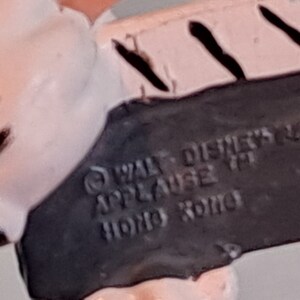 May include: Close-up of a vintage Disney figurine base. The black base features the text "WALT DISNEY'S" and "APPLAUSE TM HONG KONG". The figurine is white and pink with black accents.