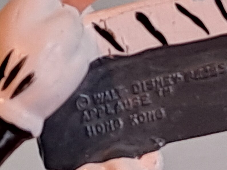 May include: Close-up of a vintage Disney figurine base. The black base features the text "WALT DISNEY'S" and "APPLAUSE TM HONG KONG". The figurine is white and pink with black accents.