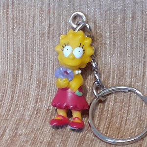 May include: Yellow cartoon character keychain with a red dress and a purple flower. The character has yellow hair, blue eyes, and a pink necklace. The keychain is attached to a silver key ring.