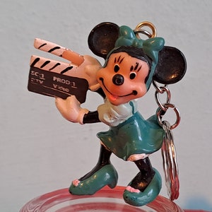 May include: A Minnie Mouse keychain figurine holding a film clapperboard. The figurine is wearing a teal dress and bow, with black ears and shoes. The clapperboard has the text "SC-1 PROD-1 Video".