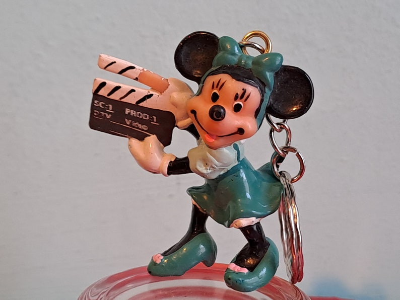 May include: A Minnie Mouse keychain figurine holding a film clapperboard. The figurine is wearing a teal dress and bow, with black ears and shoes. The clapperboard has the text "SC-1 PROD-1 Video".
