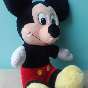 May include: A plush Mickey Mouse toy with black ears, a black and red outfit, and yellow shoes. The toy is sitting on a white surface.