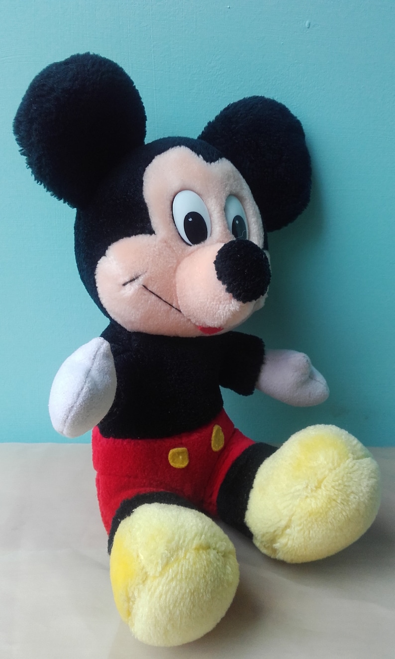May include: A plush Mickey Mouse toy with black ears, a black and red outfit, and yellow shoes. The toy is sitting on a white surface.