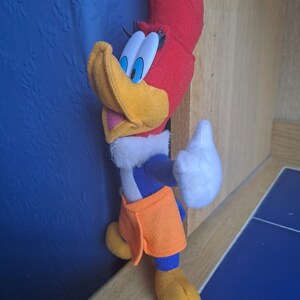 May include: A plush toy of Woody Woodpecker, a cartoon character, with a red head, yellow beak, blue and white shirt, and orange pants. The toy is standing on a wooden surface.