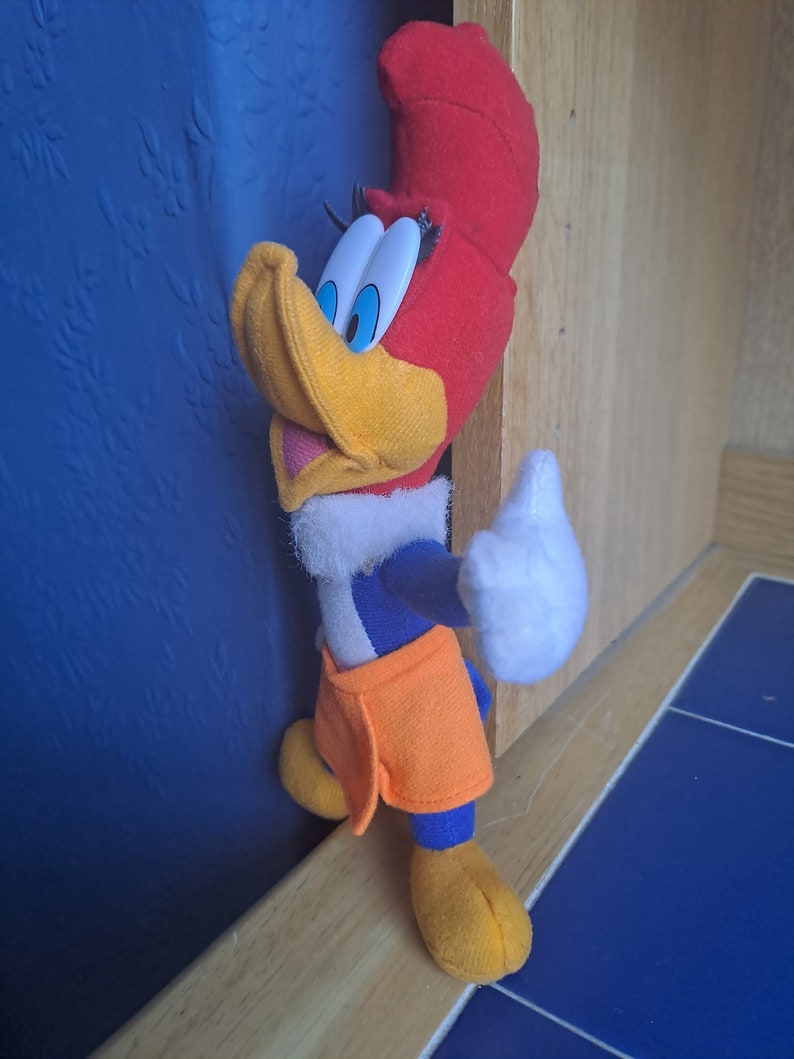 May include: A plush toy of Woody Woodpecker, a cartoon character, with a red head, yellow beak, blue and white shirt, and orange pants. The toy is standing on a wooden surface.