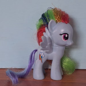 Puede incluir: Un juguete My Little Pony azul con una melena y cola arcoíris. El pony tiene un ojo rosa y morado y un símbolo de arcoíris en su flanco.