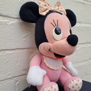 May include: A plush Minnie Mouse doll in pink, black, and white. The doll has large black ears, a pink face, and a polka-dot bow. It is wearing a white bib and has polka-dot accents on its feet.