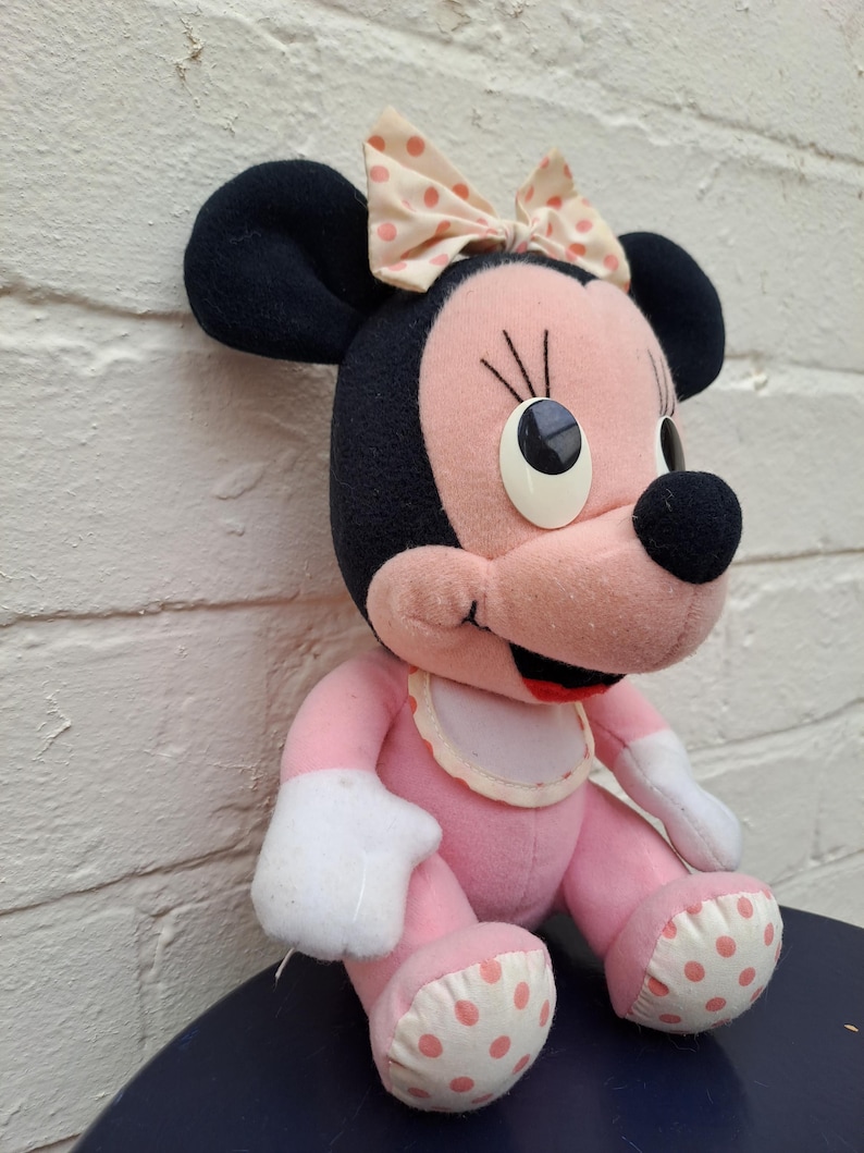 May include: A plush Minnie Mouse doll in pink, black, and white. The doll has large black ears, a pink face, and a polka-dot bow. It is wearing a white bib and has polka-dot accents on its feet.