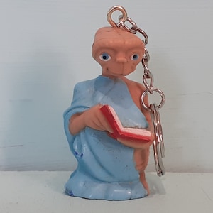 May include: A blue and red plastic keychain featuring a figurine of an alien holding a book. The alien has large eyes and a light brown skin tone.